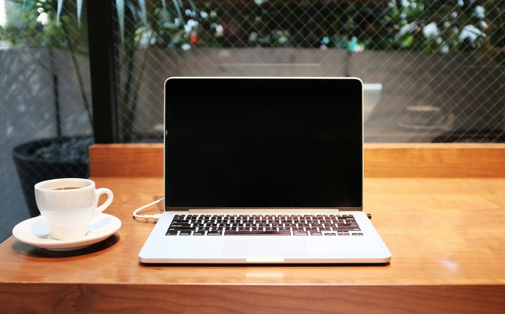 Una laptop abierta con el cargador enchufado y un café chico, sobre una mesa de madera junto a la ventana.